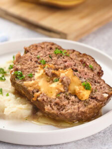 A slice of stuffed meatloaf with melted cheese on top, served on a white plate with mashed potatoes.