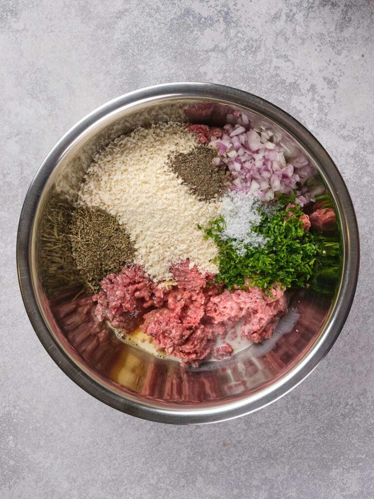 A metal bowl containing raw ground meat, breadcrumbs, chopped onions, herbs, and seasoning on a gray surface.