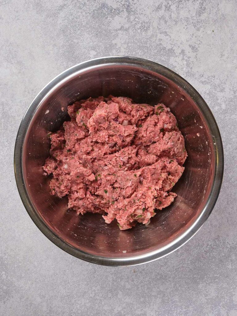 Ground beef mixed with herbs in a stainless steel bowl on a gray textured surface.