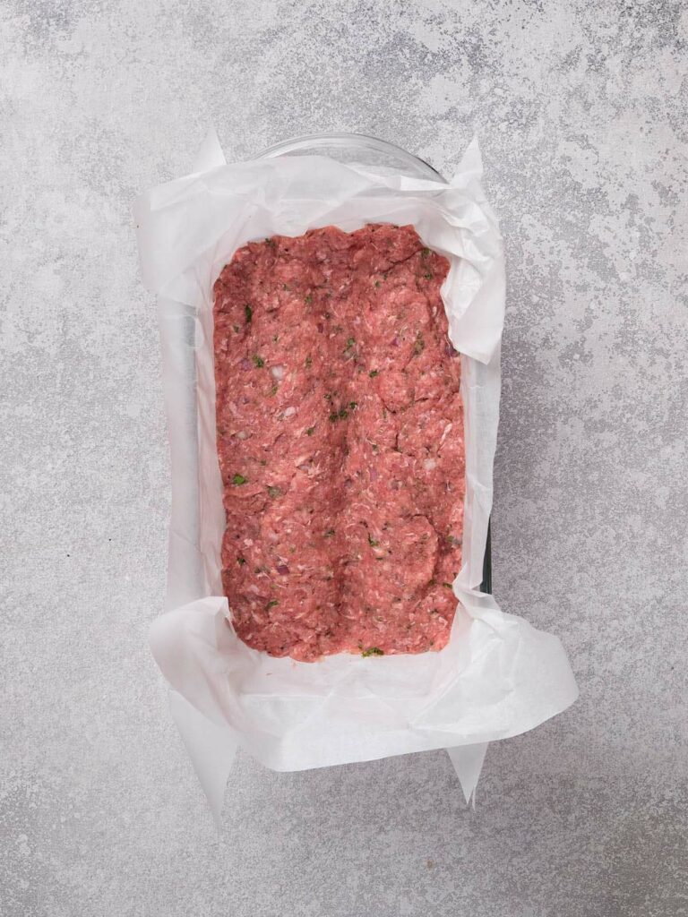 Raw ground meat mixture in a parchment-lined loaf pan on a gray surface.