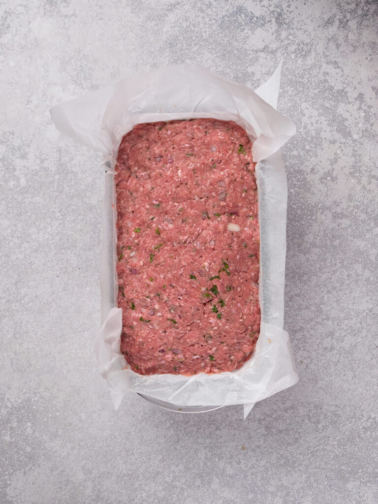 A loaf pan filled with uncooked meatloaf mixture, topped with chopped herbs, on a gray surface.