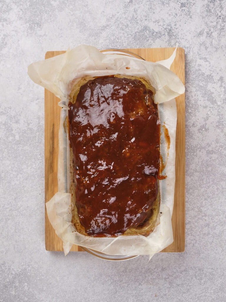 A glazed meatloaf on parchment paper in a rectangular baking dish atop a wooden board.