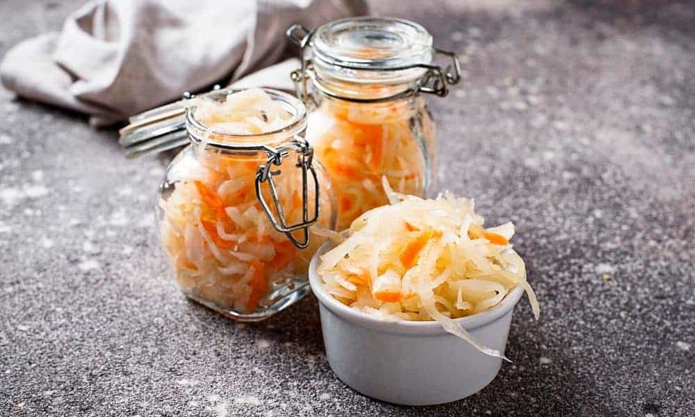 Homemade sauerkraut in a glass jar and served in a small white bowl on a kitchen countertop.