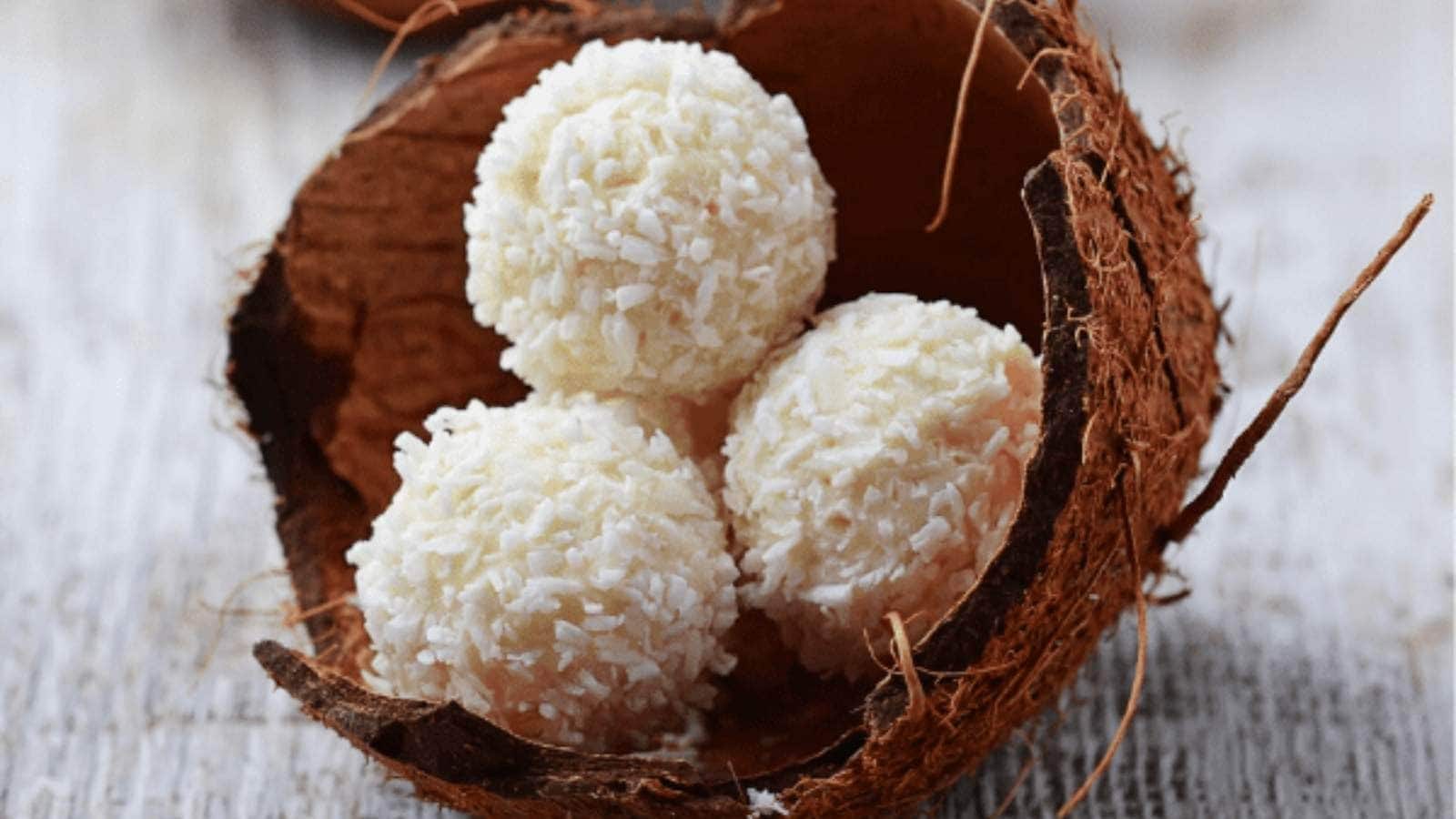 Three coconut-covered macaroons are placed inside a halved coconut shell on a light wooden surface.