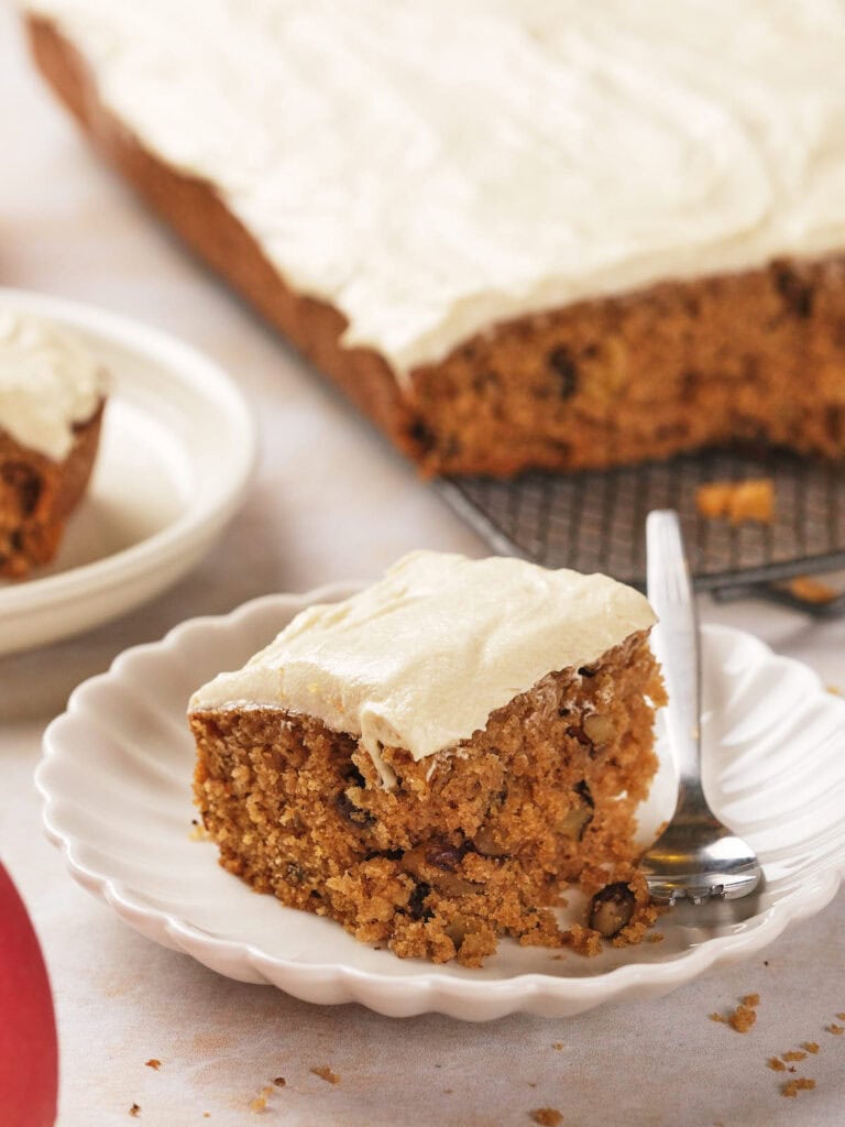 A slice of frosted applesauce cake with nuts is on a white plate with a fork.