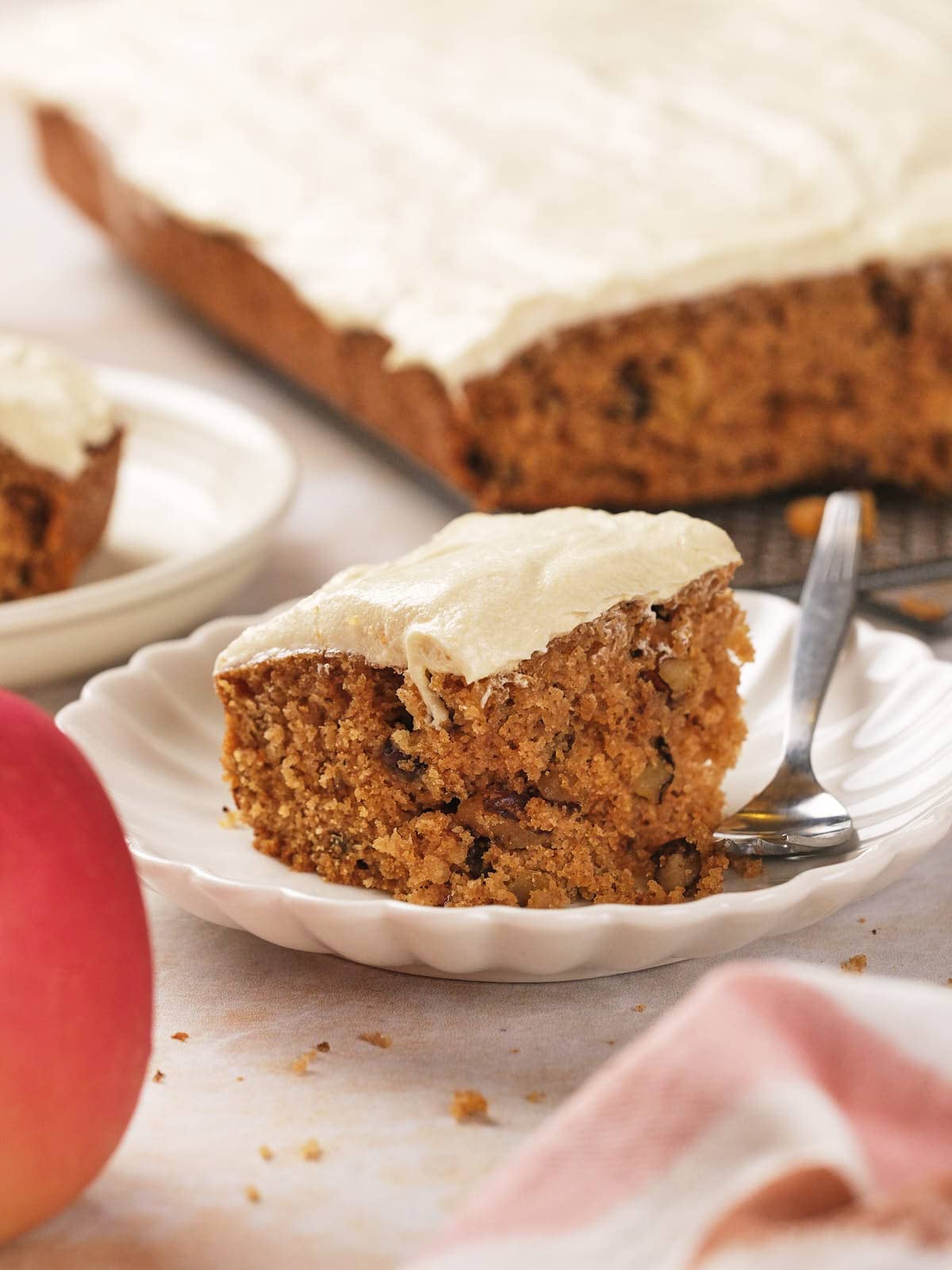 A slice of frosted applesauce cake with visible nuts on a white plate, accompanied by a silver fork. The whole cake is partially seen in the background.