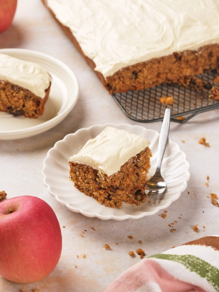 A slice of frosted applesauce on a plate with a fork. The rest of the cake is on a cooling rack. An apple is placed nearby.