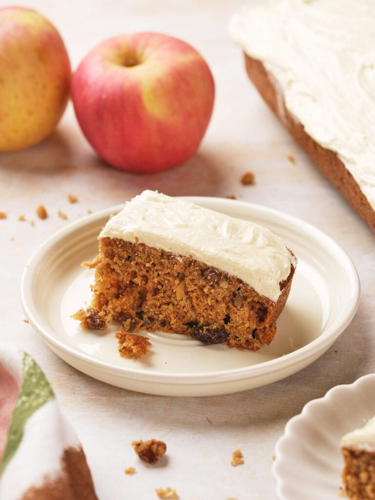 A slice of frosted applesauce cake on a white plate, surrounded by two apples and the rest of the cake in the background.