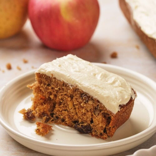 A slice of frosted applesauce cake on a white plate with two whole apples in the background.