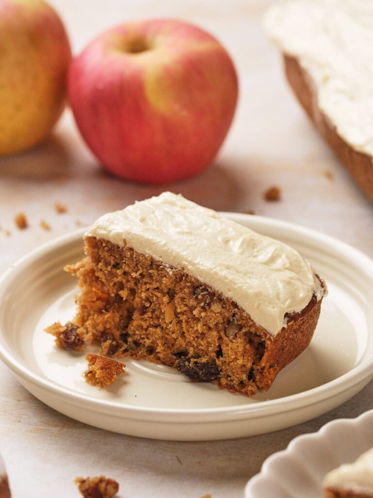 A slice of frosted applesauce cake on a white plate with two whole apples in the background.