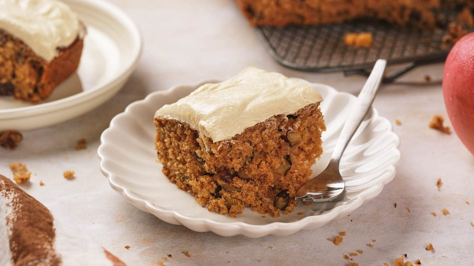 A slice of frosted applesauce cake with nuts on a white scalloped plate, accompanied by a fork.