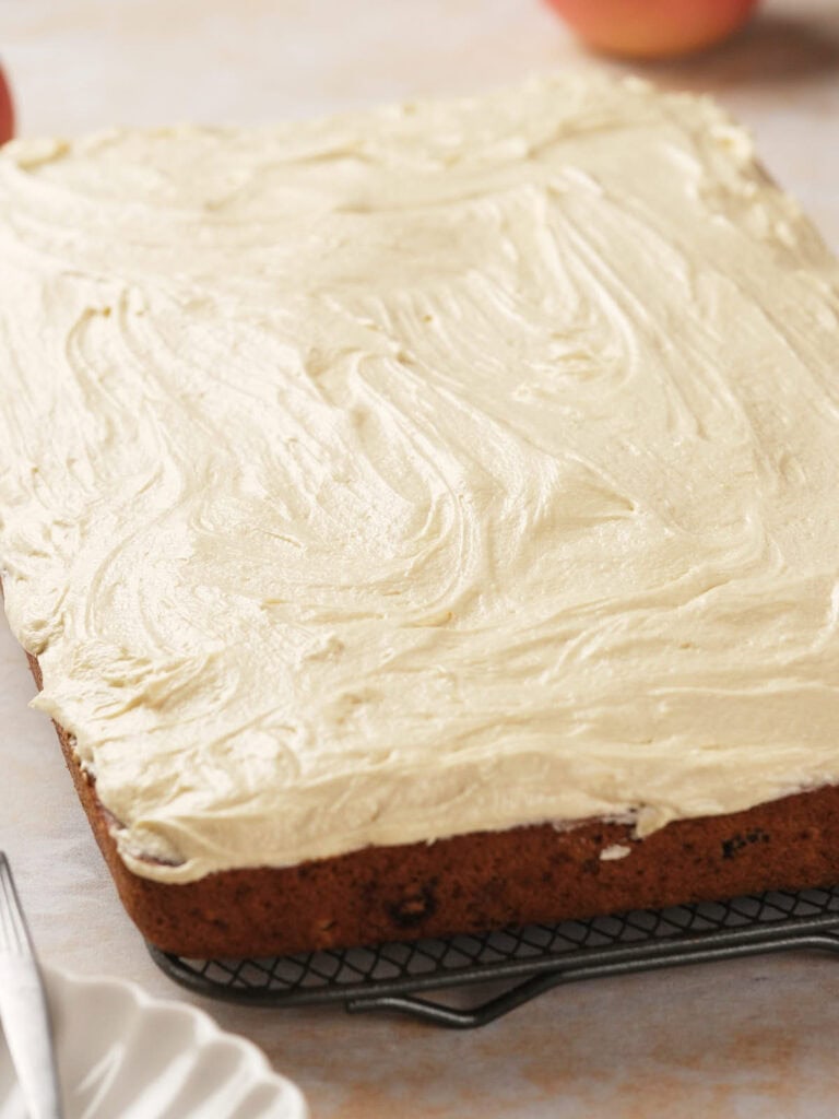 Rectangular apple cake with cream cheese frosting on a cooling rack, with part of a fork and plate visible nearby.