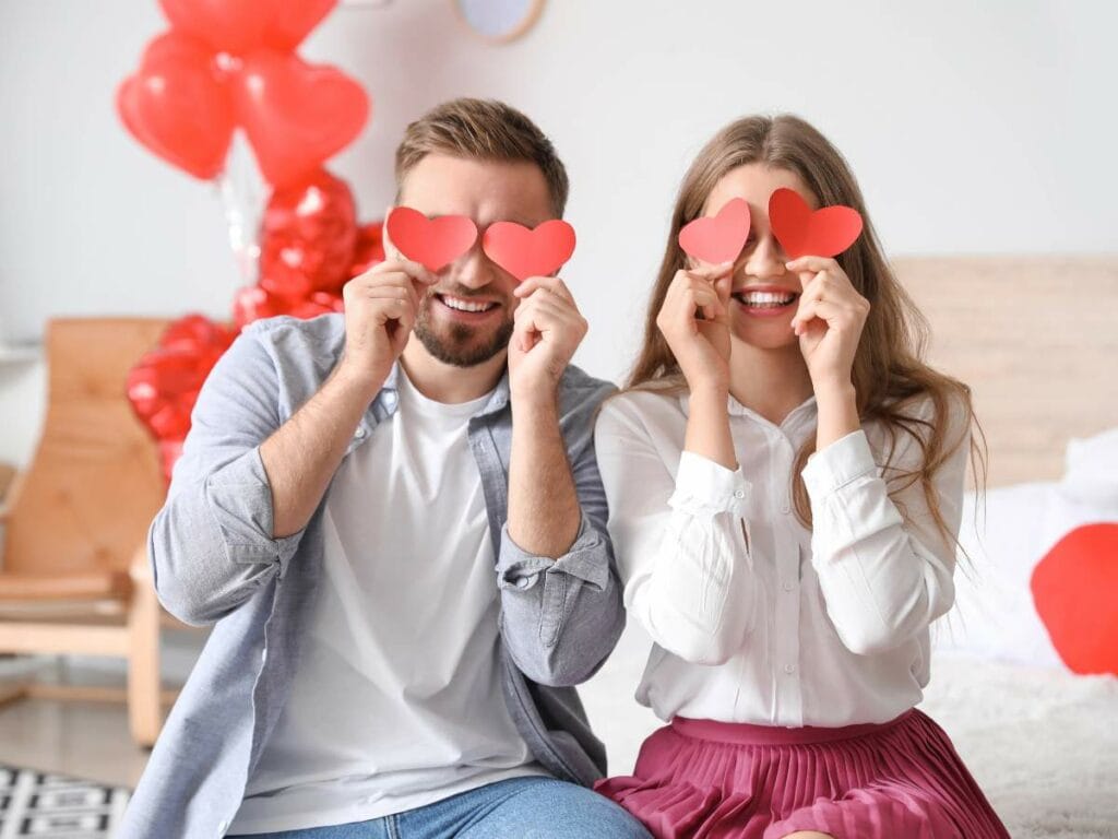 A man and woman sitting indoors hold red paper hearts over their eyes, smiling.