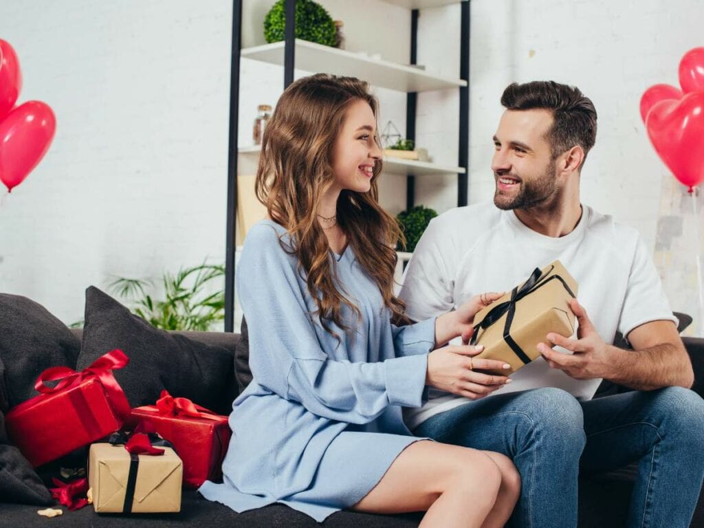 A couple sits on a couch, smiling at each other while exchanging gifts.