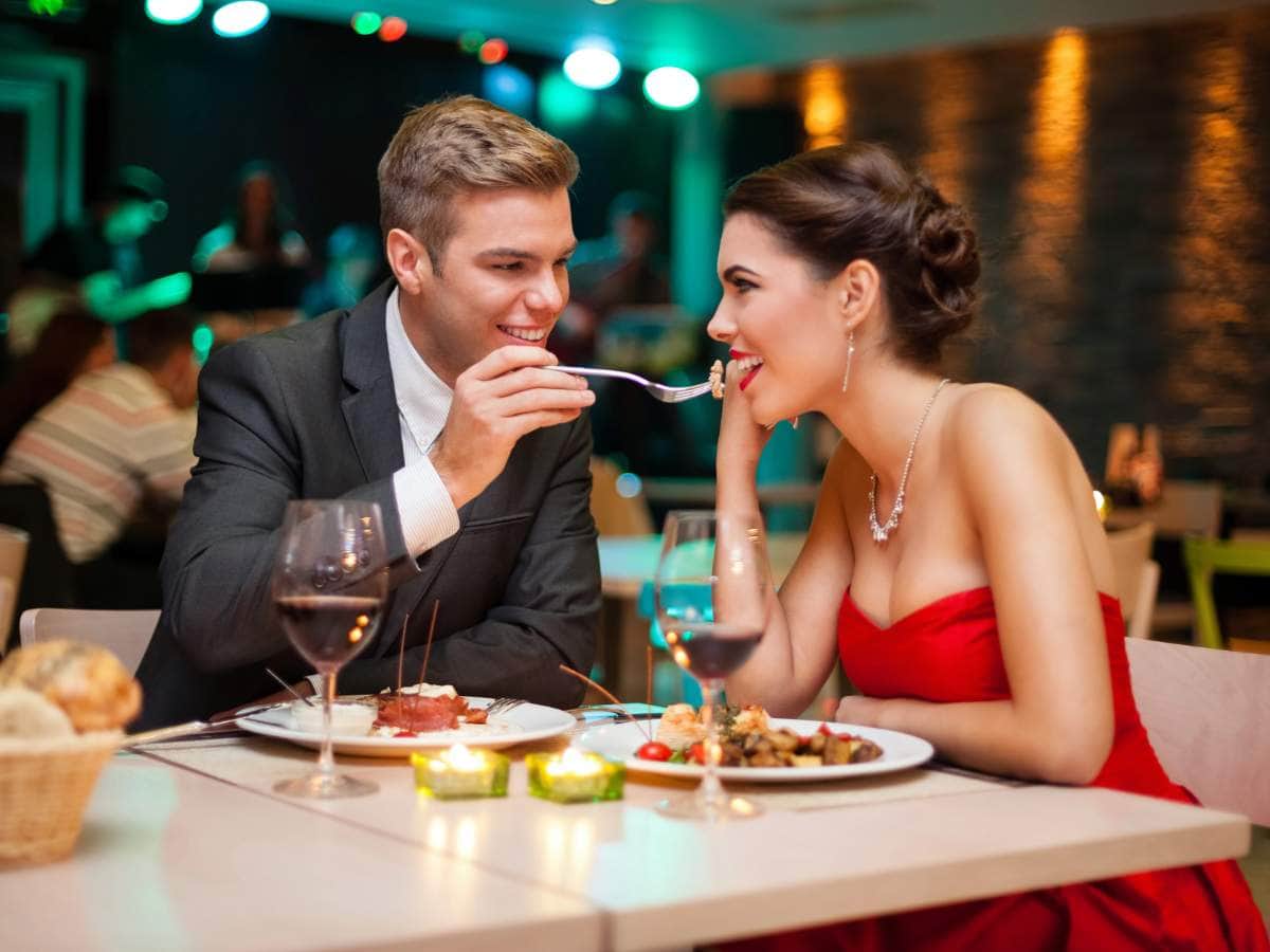 A man in a suit feeds a woman in a red dress at a restaurant.