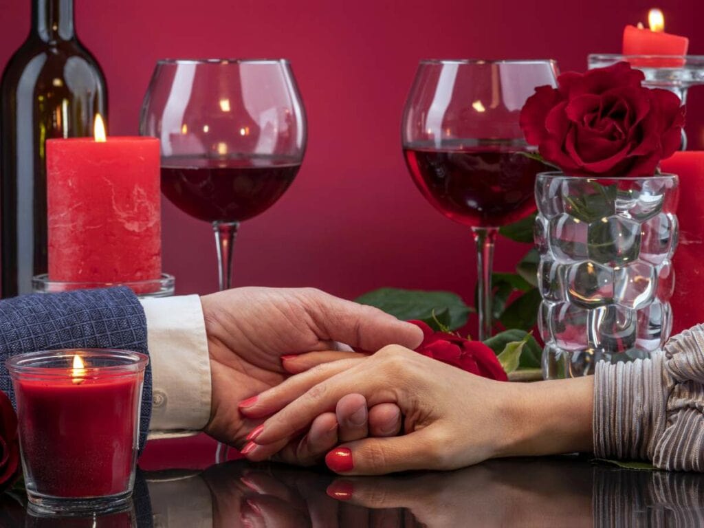 Two people holding hands across a table set with red candles, red wine glasses, and red roses.