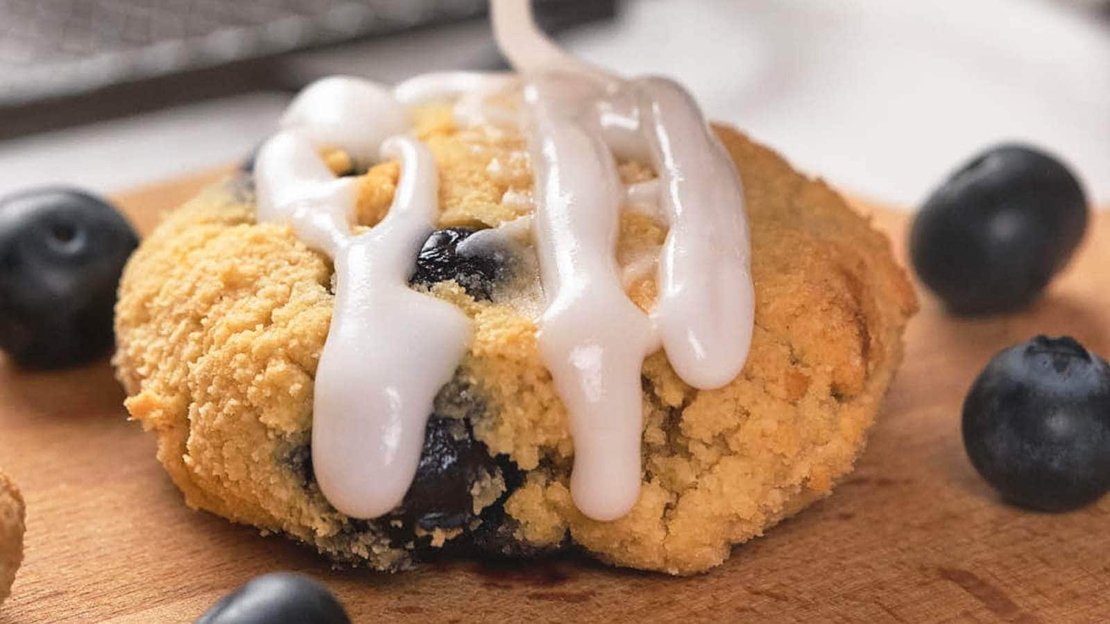 Close-up of a blueberry scone drizzled with white icing, surrounded by fresh blueberries on a wooden surface.