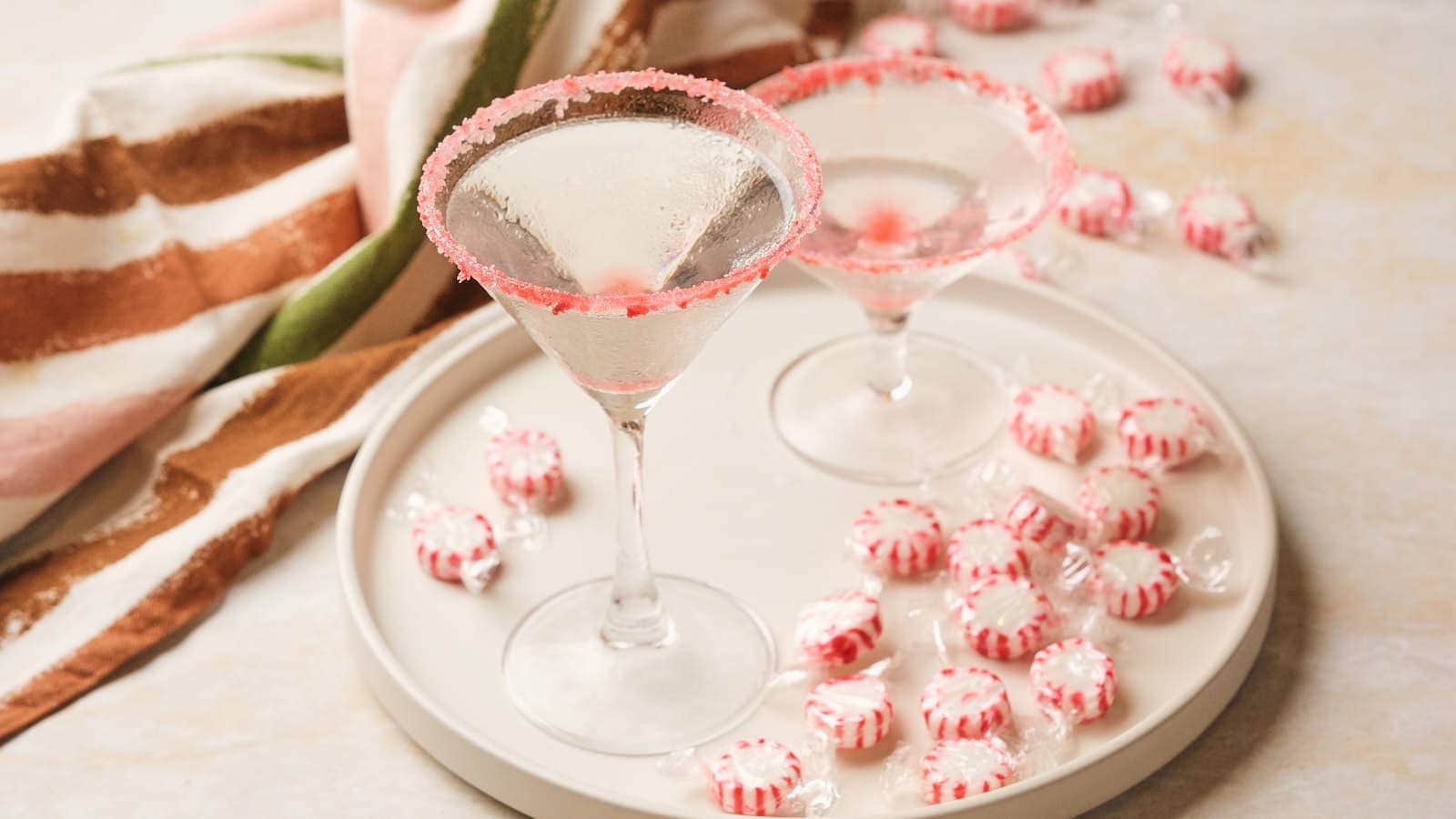 Two martini glasses with pink sugar rims on a tray, surrounded by peppermint candies and a striped cloth.