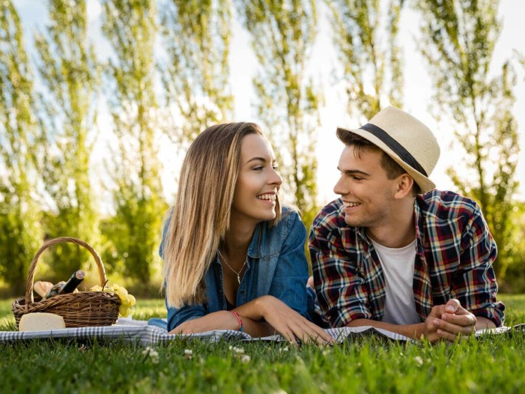 A couple lies on a picnic blanket on grass, smiling at each other.