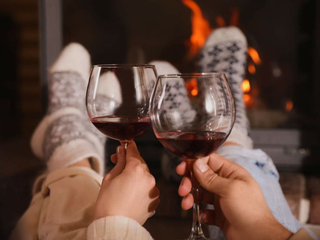 Two people clink wine glasses in front of a fireplace, with sock-covered feet resting nearby.