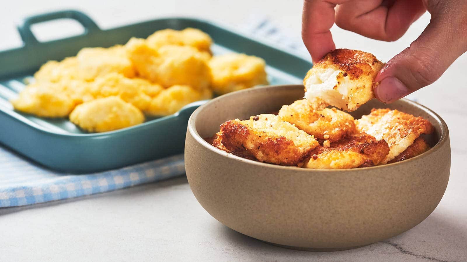 A hand picks up a piece of fried cheese curd from a bowl.