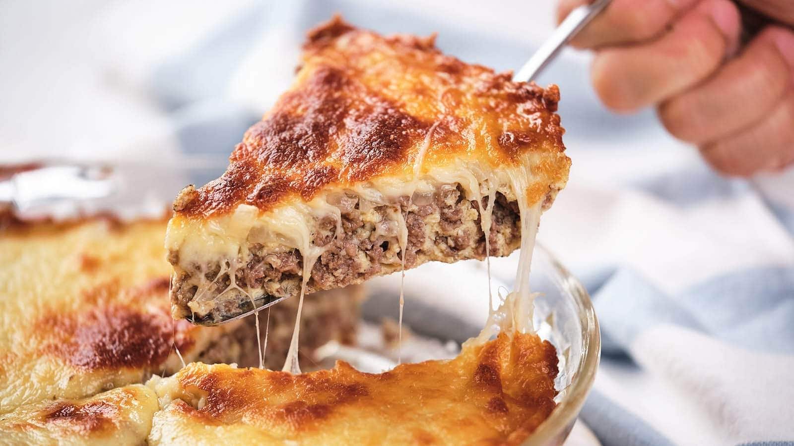 A hand lifts a cheesy slice of cheeseburger pie, showing melted cheese and a golden-brown top.