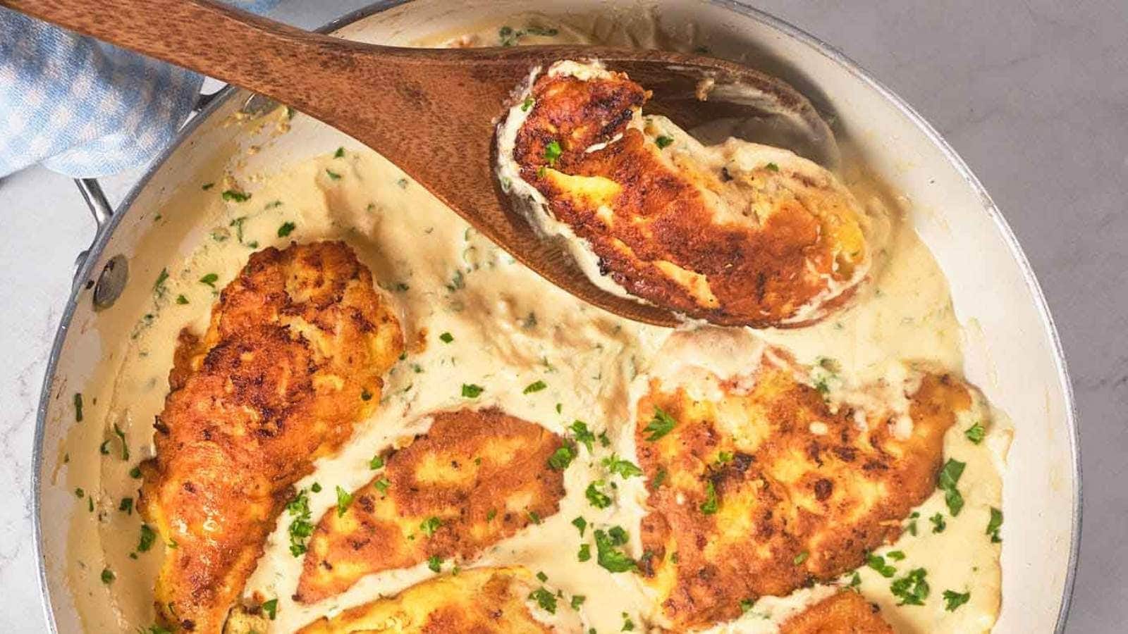 A skillet filled with chicken francese garnished with chopped herbs, being stirred with a wooden spoon.