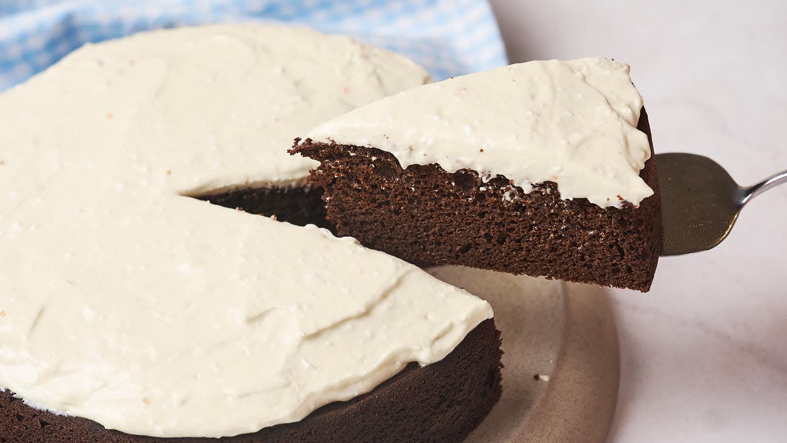 A slice of chocolate cake with white frosting being lifted from the whole cake on a light-colored platter.