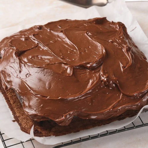 Square chocolate fudge cake with chocolate frosting on a cooling rack, parchment paper underneath, and a cake server nearby.