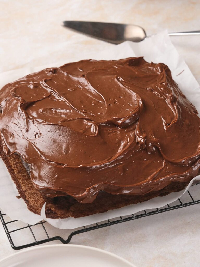 Square chocolate fudge cake with chocolate frosting on a cooling rack, parchment paper underneath, and a cake server nearby.