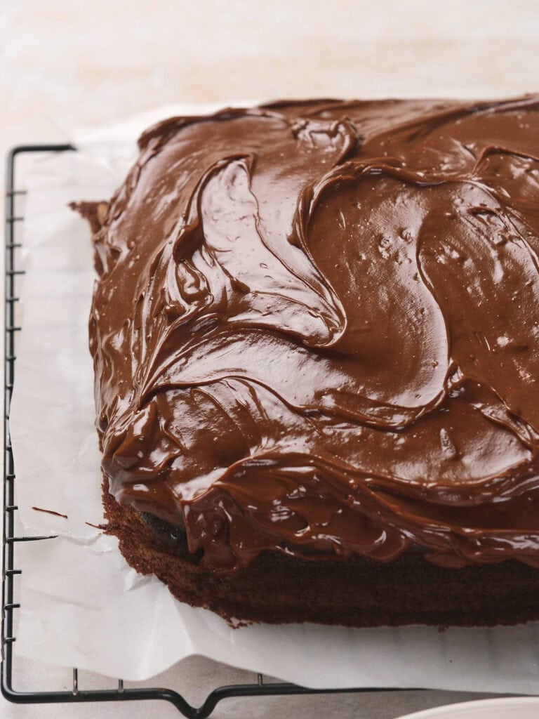 A square chocolate fudge cake with smooth, glossy chocolate frosting rests on parchment paper over a cooling rack.