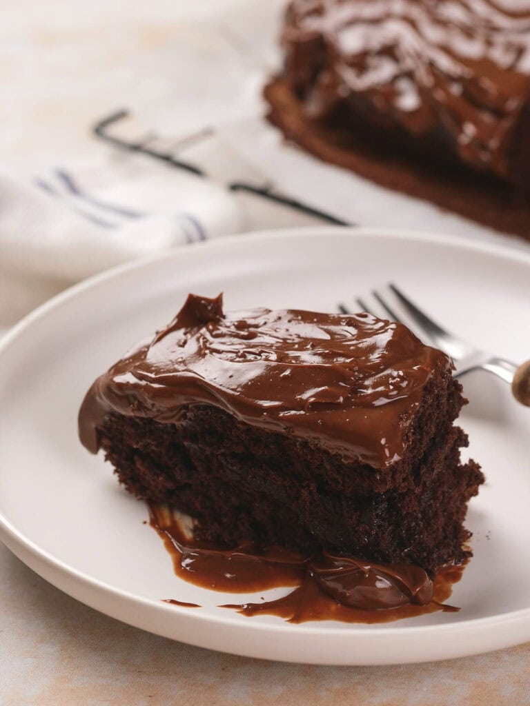 A slice of chocolate fudge cake with glossy frosting on a white plate, with a fork beside it.