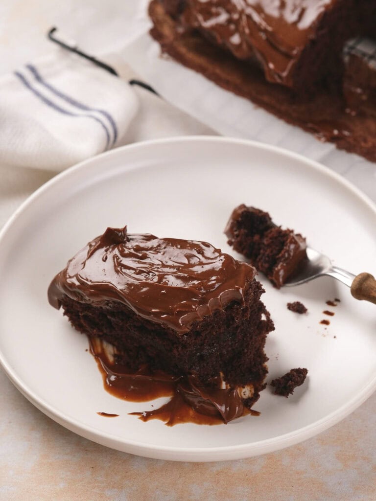 A slice of chocolate fudge cake with glossy frosting is on a white plate.