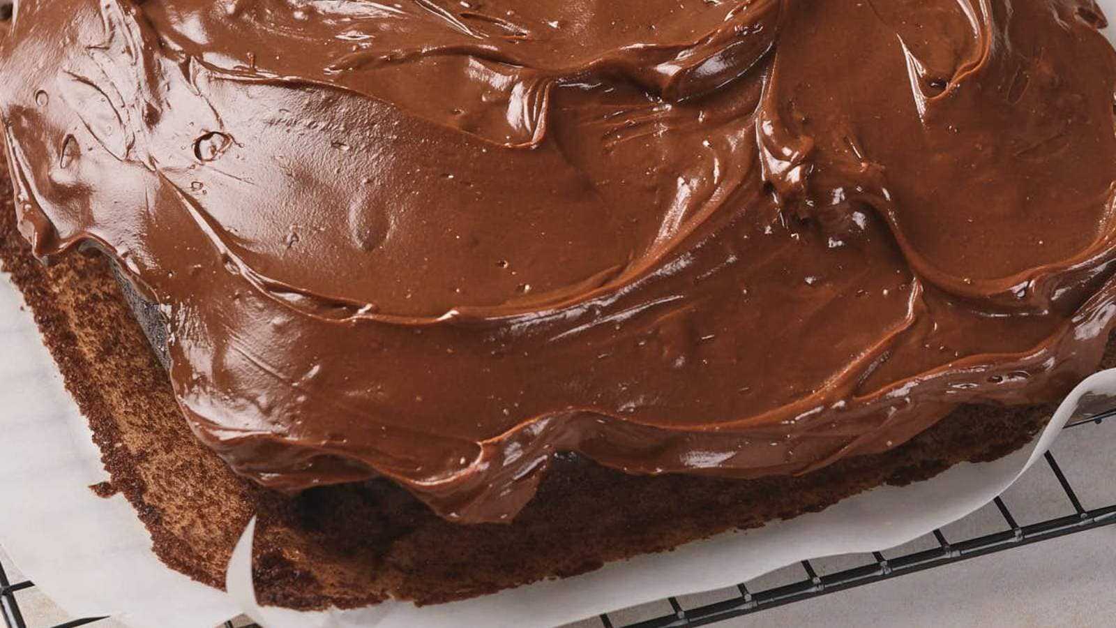 A close-up of a chocolate-frosted cake on a cooling rack with parchment paper underneath.