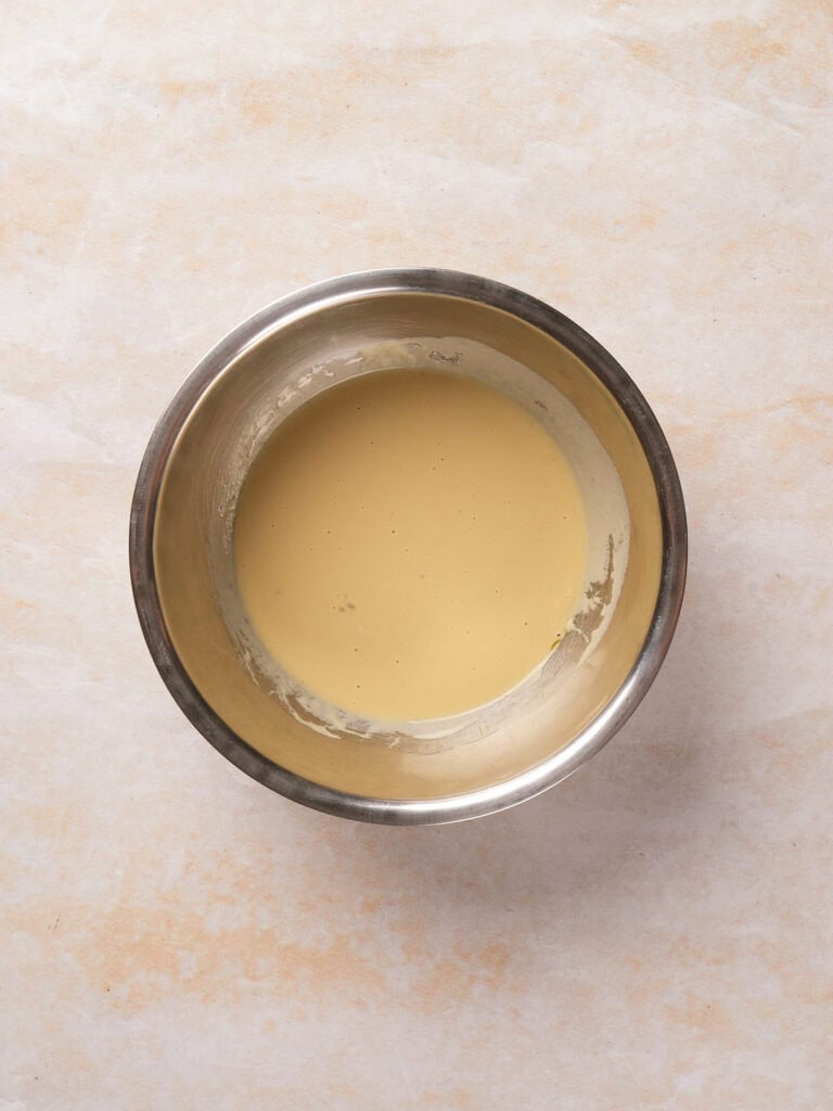 A metal bowl containing a light beige batter on a light textured surface.