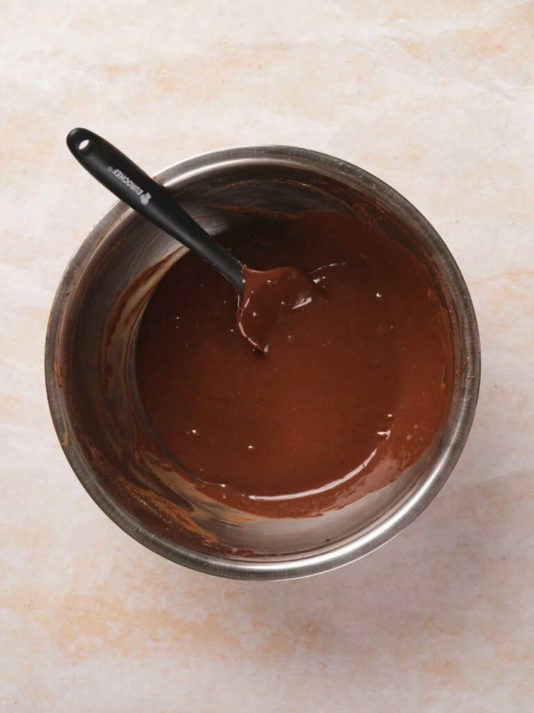 A metal bowl filled with smooth chocolate cake batter and a black-handled spatula on a light textured surface.