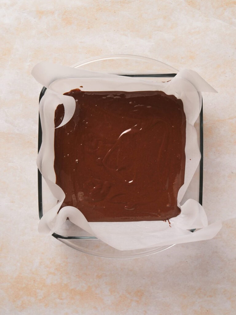 Brownie batter spread evenly in a parchment-lined square baking dish, ready for baking.