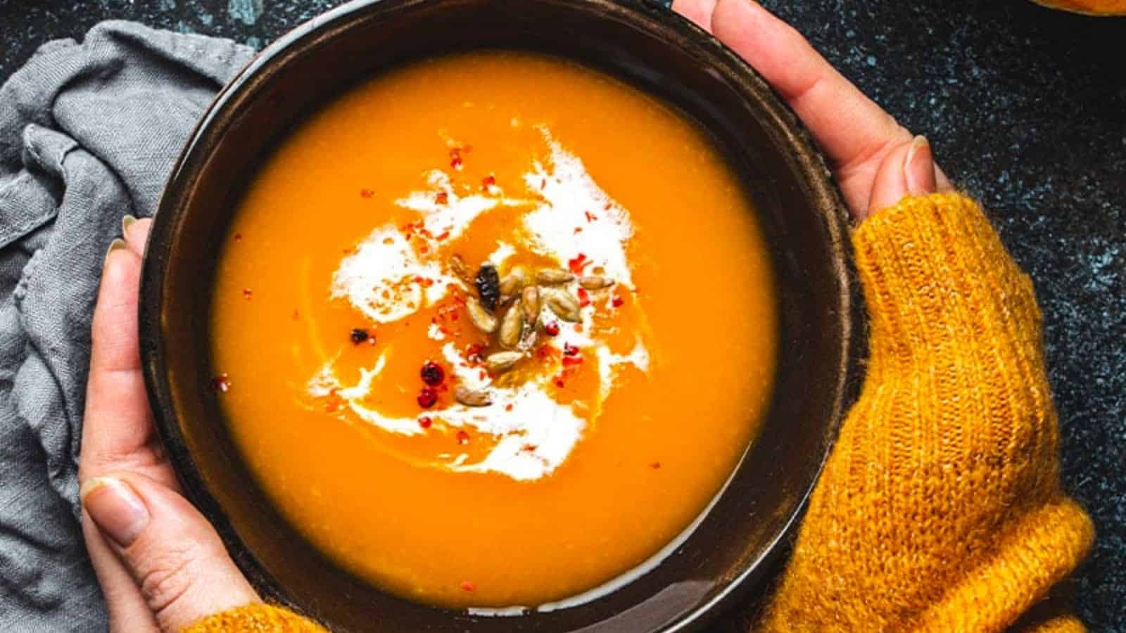 A bowl of coconut pumpkin soup garnished with cream, seeds, and spices is held by two hands in yellow sweaters.