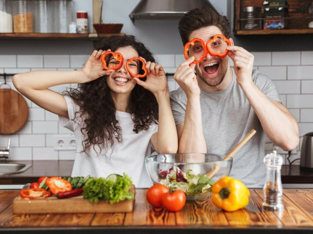 Two people in a kitchen hold sliced bell peppers over their eyes, smiling.