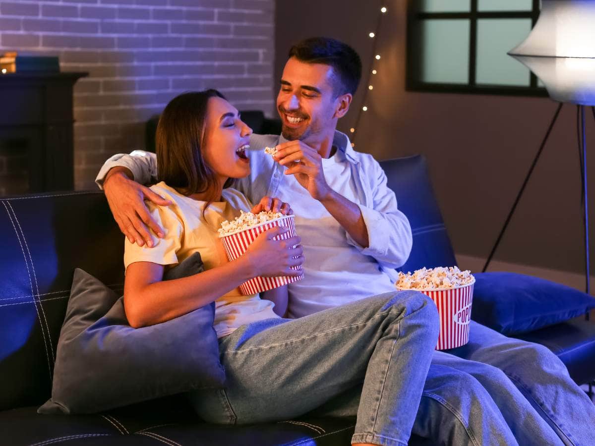 A couple sits on a sofa, sharing popcorn and smiling.