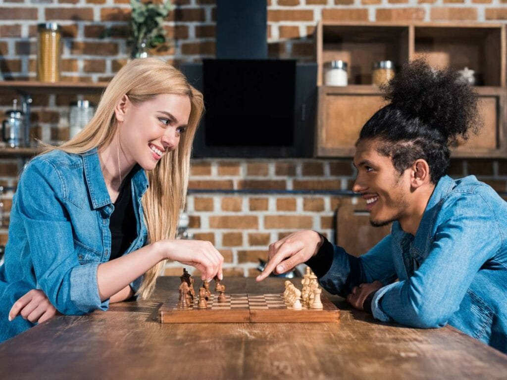 Two people playing chess at a table in a kitchen. Both are smiling and wearing denim jackets.