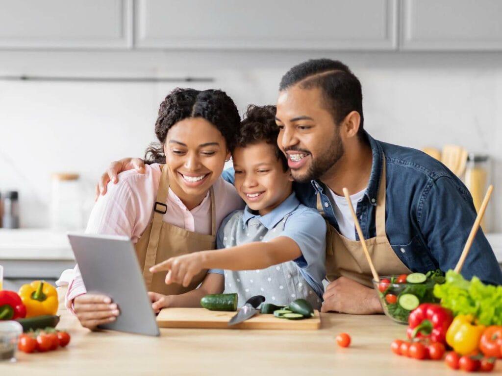 A family of three in a kitchen, wearing aprons, looking at a tablet. Various vegetables are on the countertop.