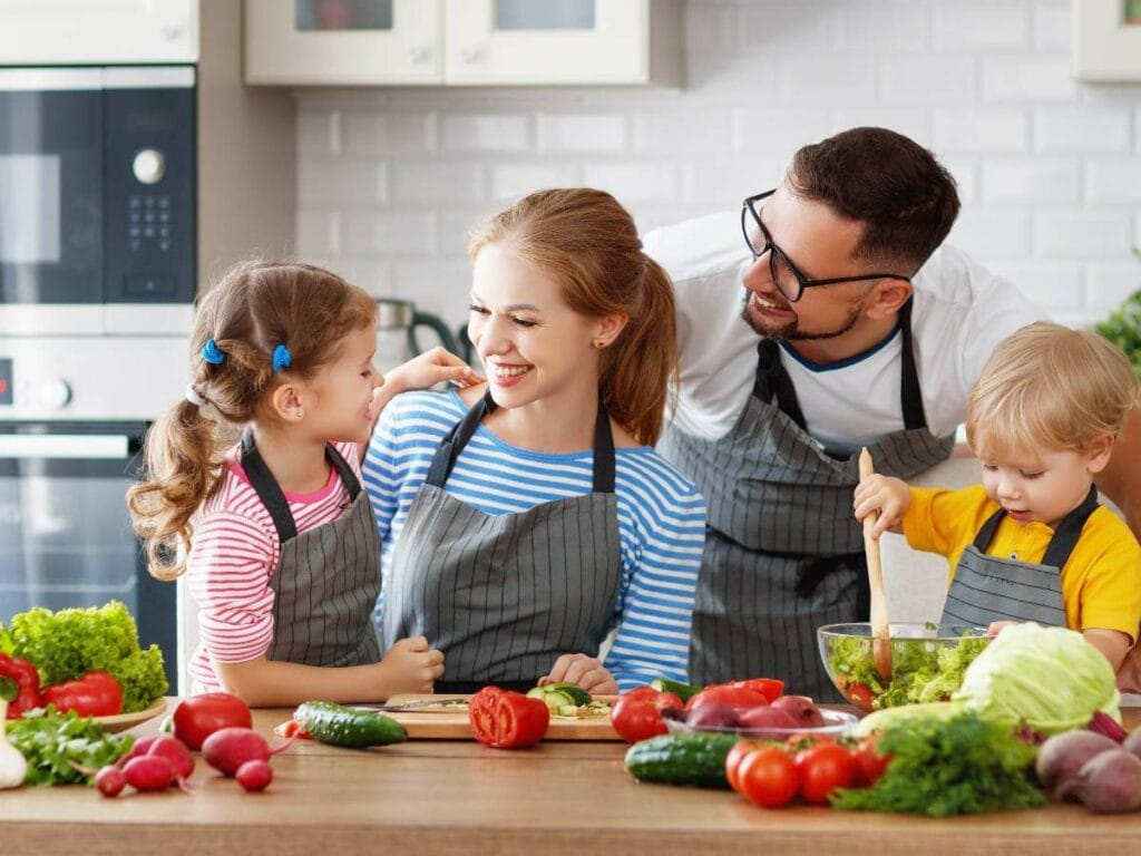 A family of four wearing aprons prepares a meal together in a kitchen.