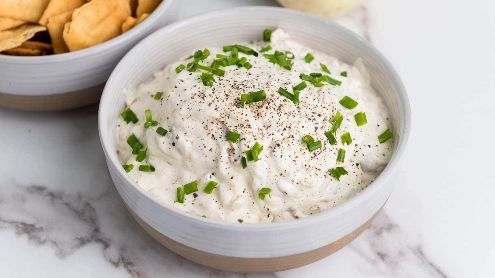 A bowl of creamy french onion dip topped with chopped chives and black pepper, placed beside a bowl of chips on a marble surface.