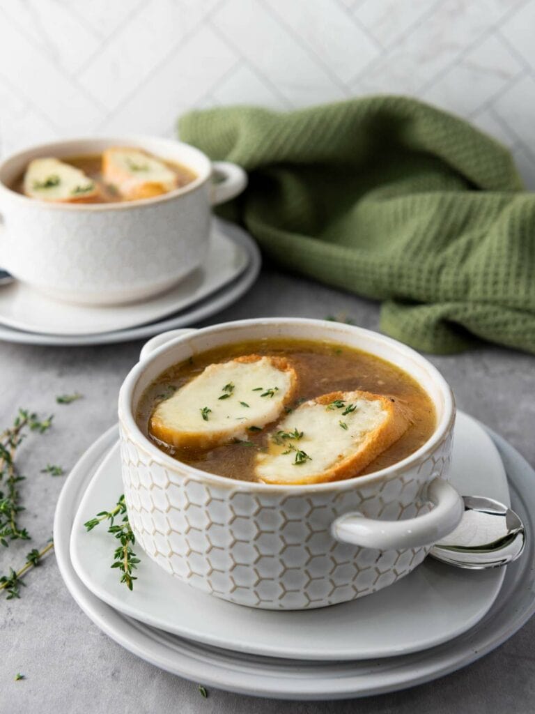 Two bowls of French onion soup topped with baguette slices and herbs.