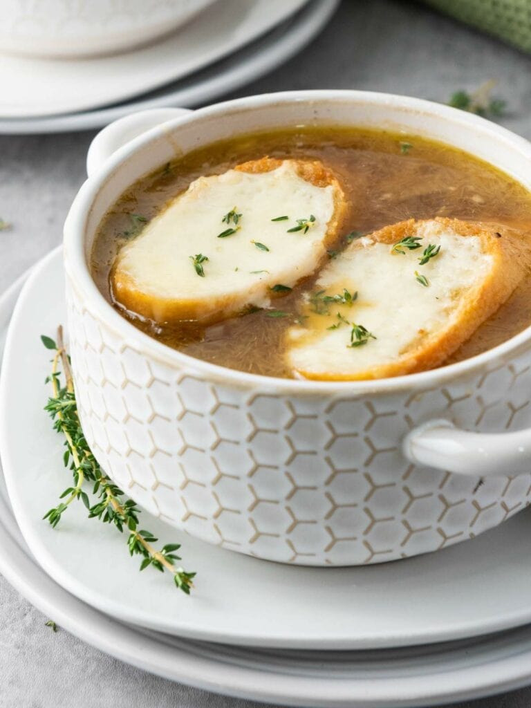 A bowl of French onion soup topped with two cheese-covered bread slices, served on a stack of white plates with a sprig of thyme.