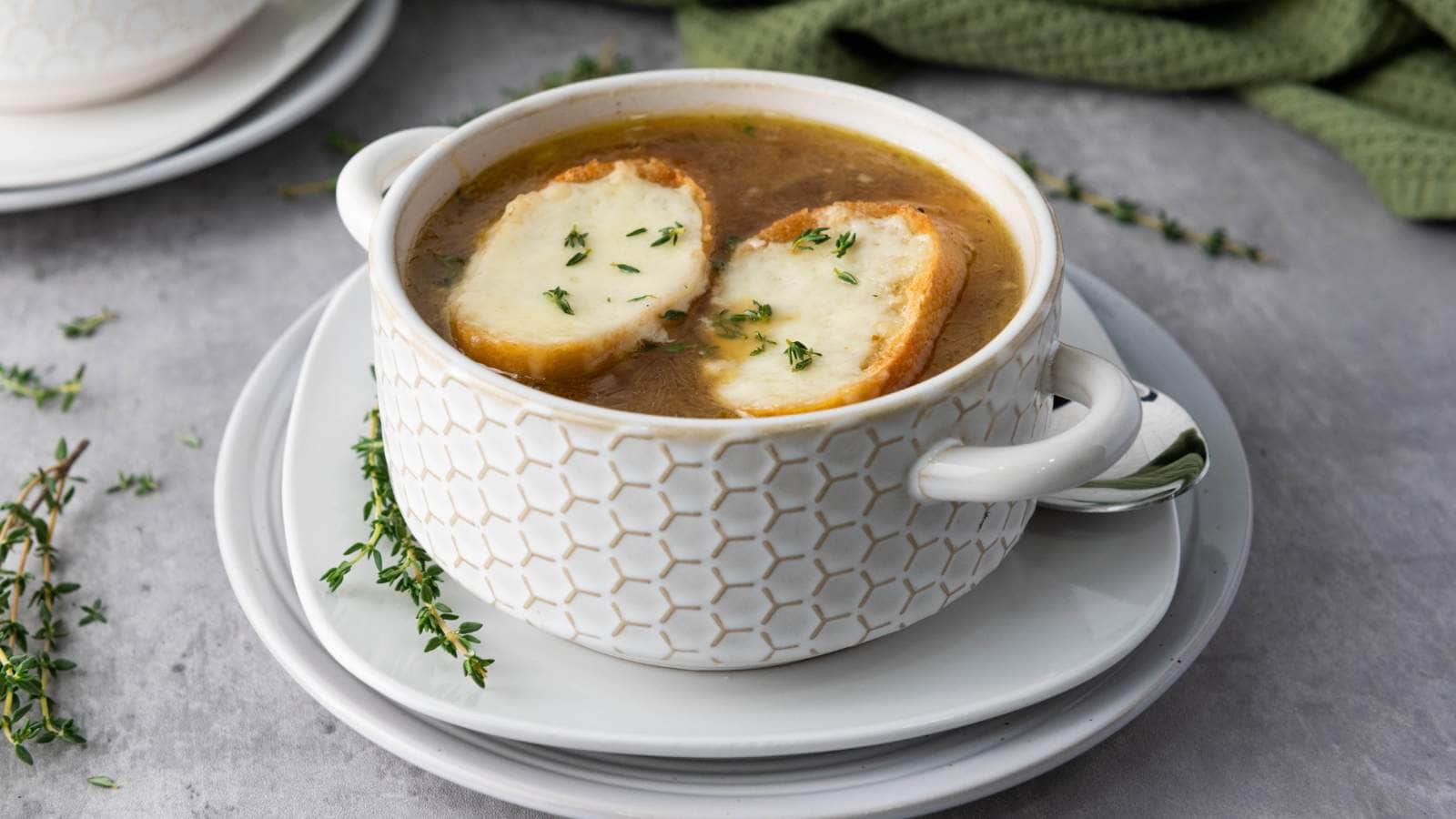 A bowl of French onion soup topped with melted cheese and herbs on bread, placed on a white plate.