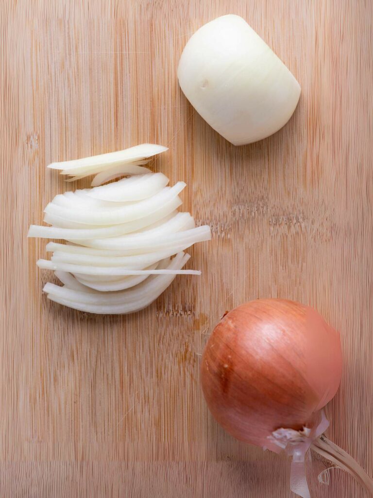 A whole brown onion, a halved peeled onion, and sliced onion are on a wooden cutting board.