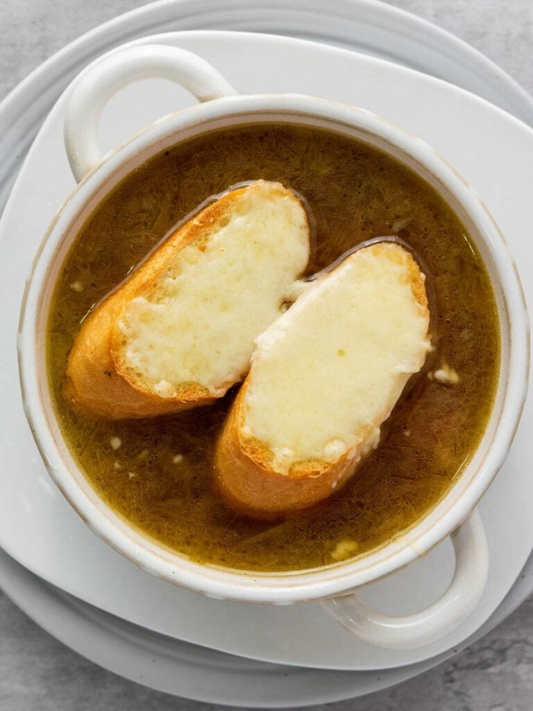 A bowl of onion soup topped with two slices of bread covered in melted cheese, served on a white plate.