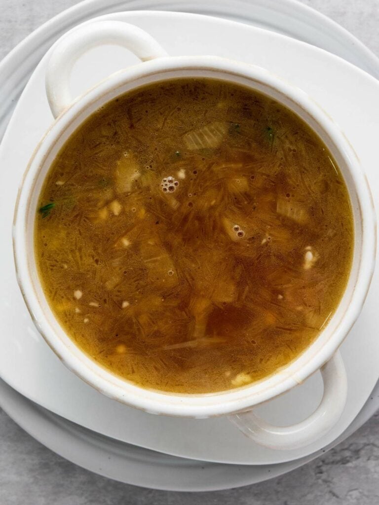 A white bowl filled with clear broth soup, placed on a matching saucer, against a light background.